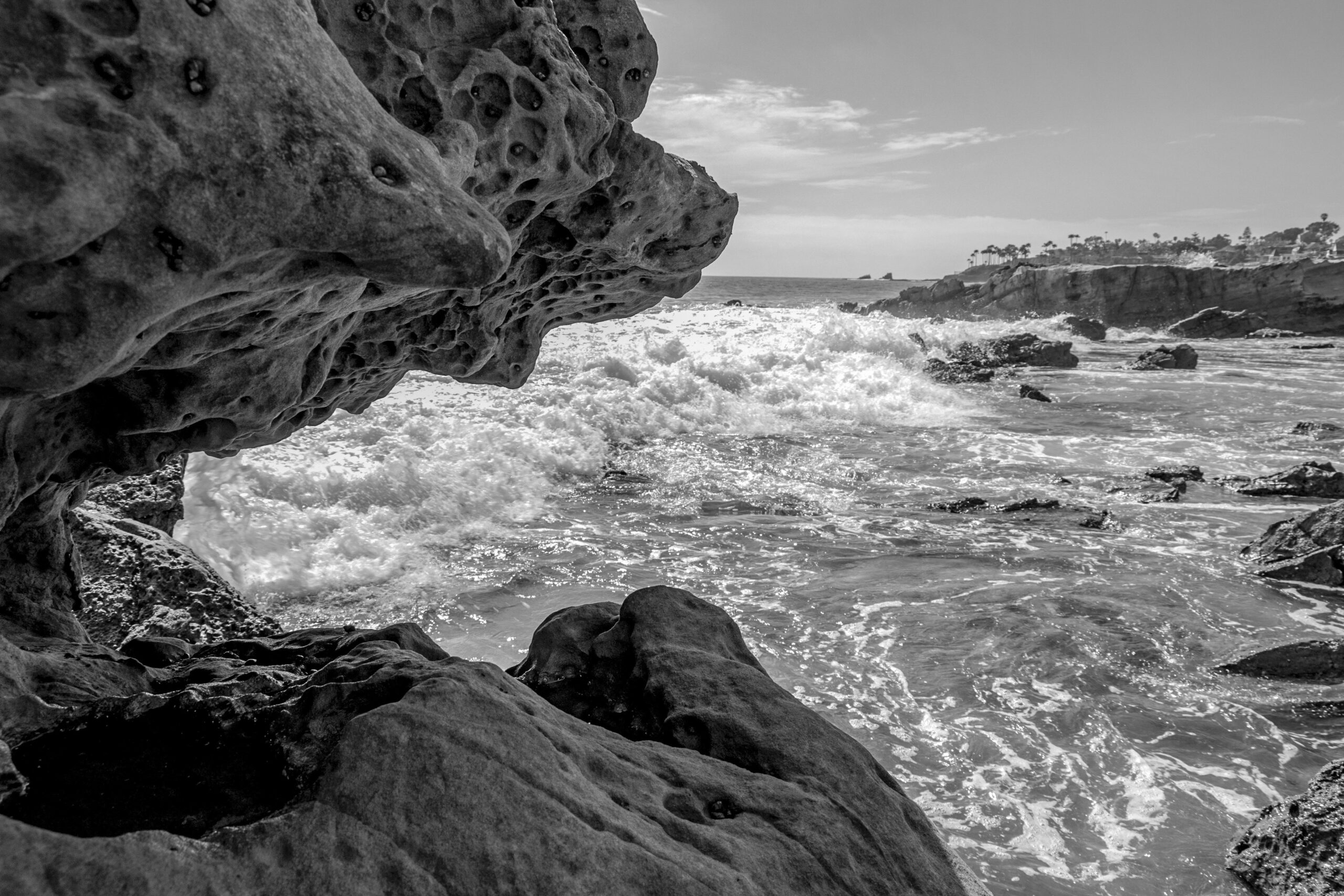 Black-and-white coastal scene framed by rock with moving water and distant shoreline.