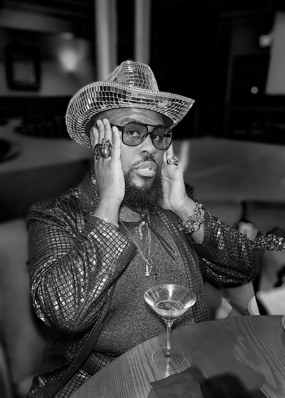 Black-and-white editorial portrait of a man in a mirrored cowboy hat and glasses seated at a table.