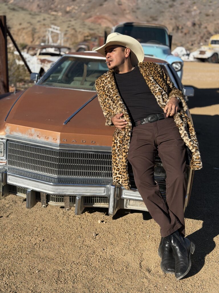 Lifestyle portrait of a man in a cowboy hat and leopard-print coat leaning against a vintage car in the desert.