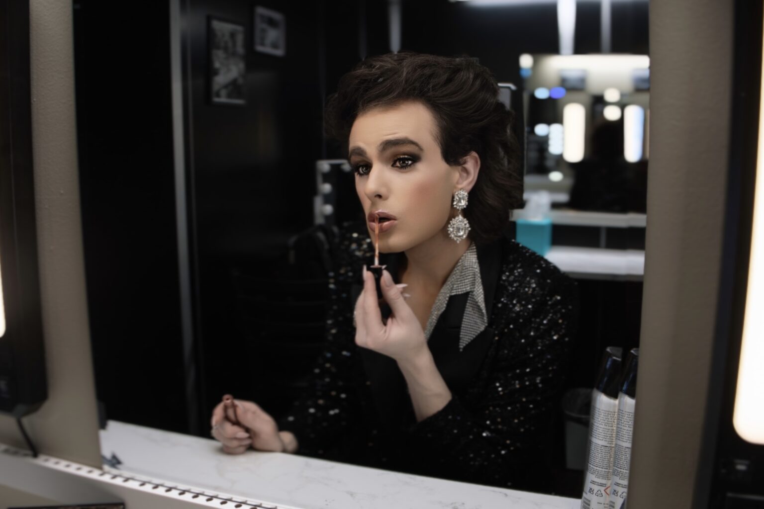 Editorial portrait of a performer applying makeup at a lighted vanity mirror.