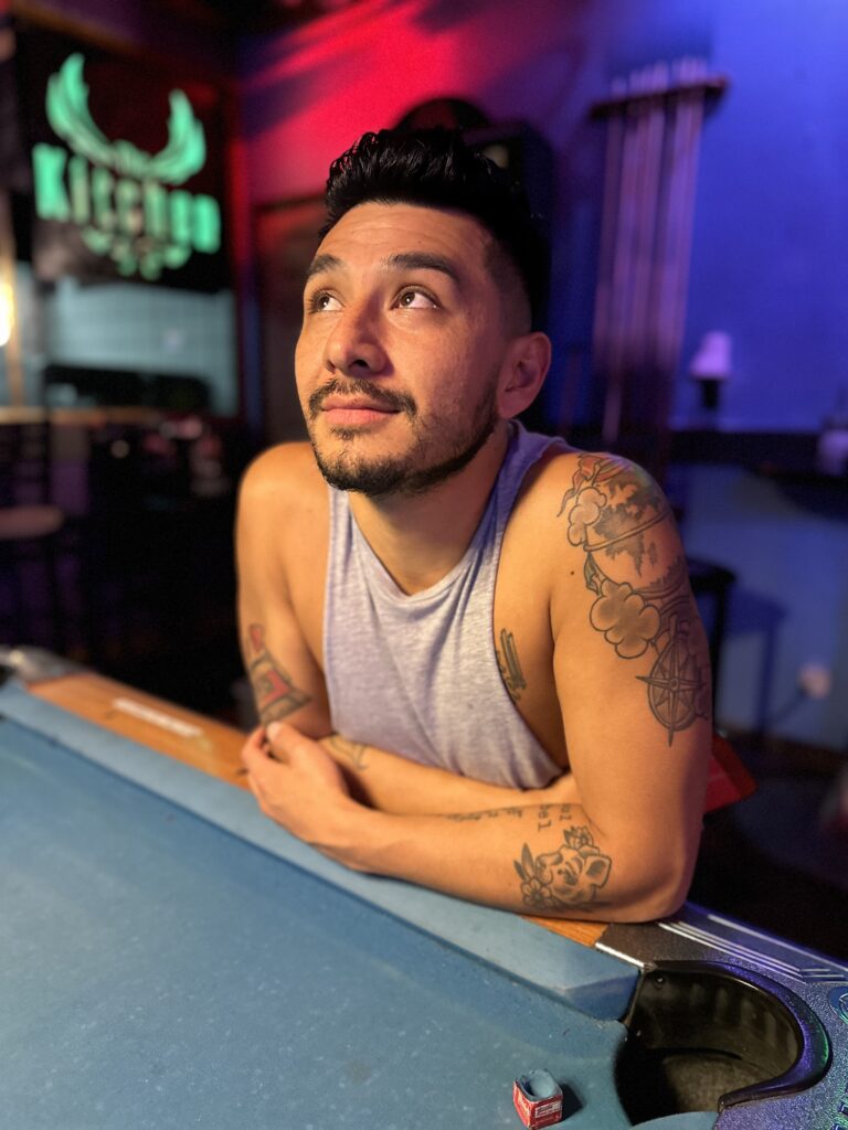Lifestyle portrait of a tattooed man leaning on a pool table in colorful ambient light.